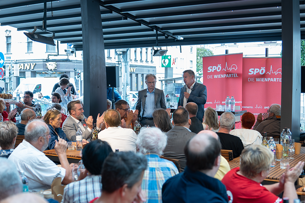 Foto (v.r.n.l.): Vizekanzler, Wohnminister Andreas Babler und Finanzminister Markus Marterbauer sprechen vor einer Gruppe von Menschen in einem Café