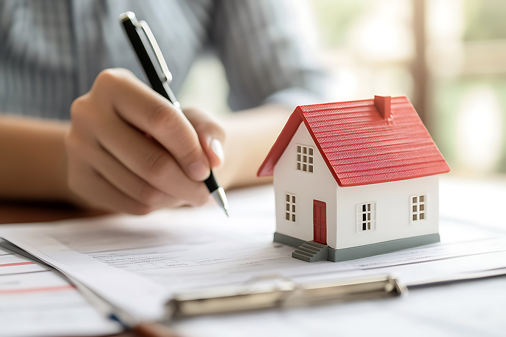 Symbolbild: Jemand hält einen Kugelschreiber in der Hand zum Unterschreiben; Auf einem Vertrag steht ein Spielzeug-Haus